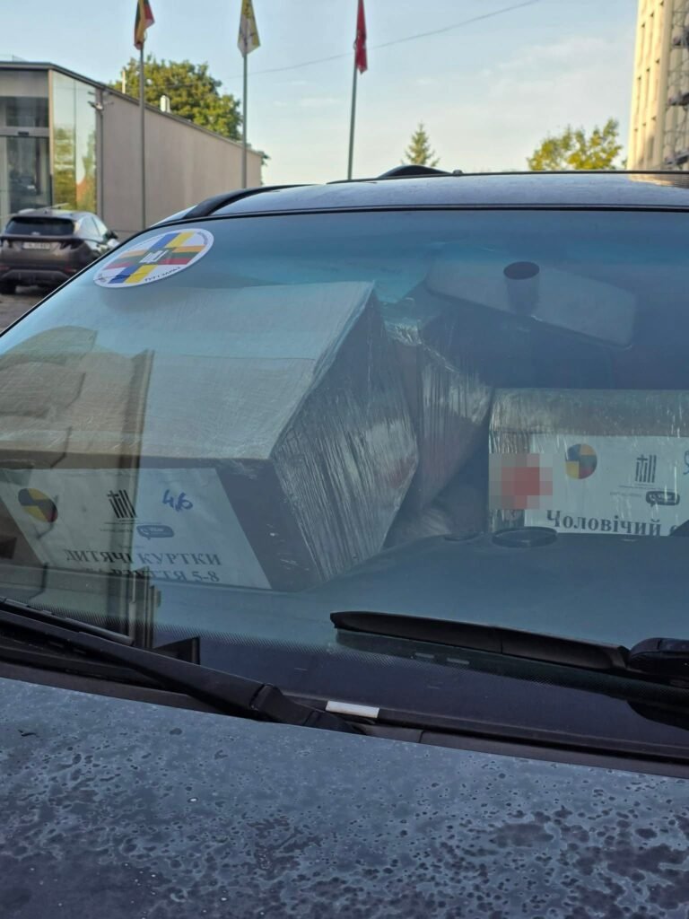 Pro automobilio priekinį stiklą matyti dėžės su drabužiais, pažymėtos „Visi išvien“ ženklu