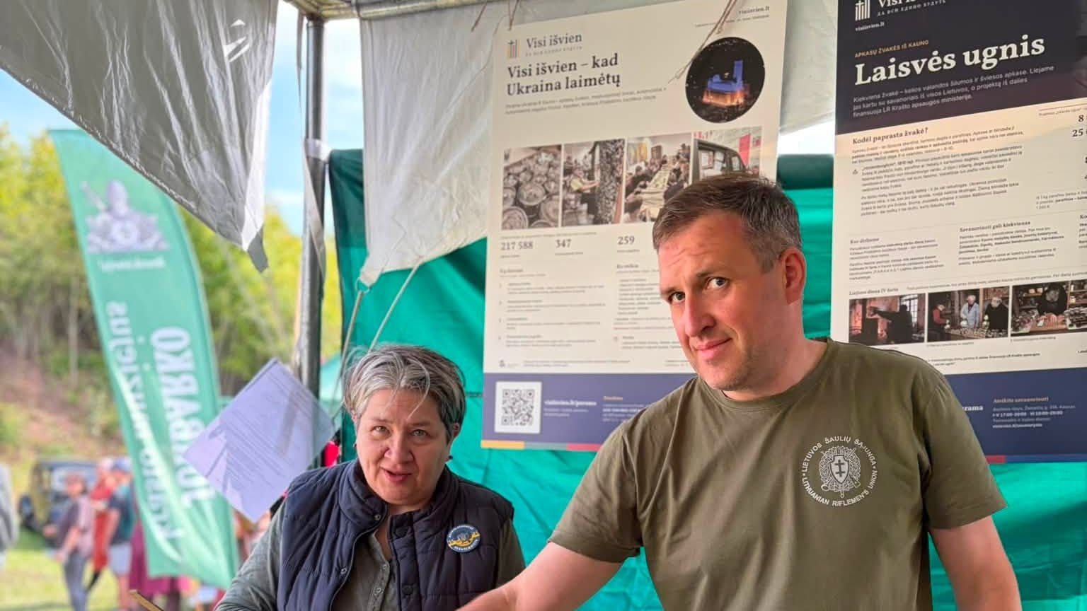 Tatjana Narkevičienė ir savanoris „Visi išvien“ palapinėje festivalyje „Fort-Unija“ IV forte, už jų – plakatai „Laisvės ugnis“