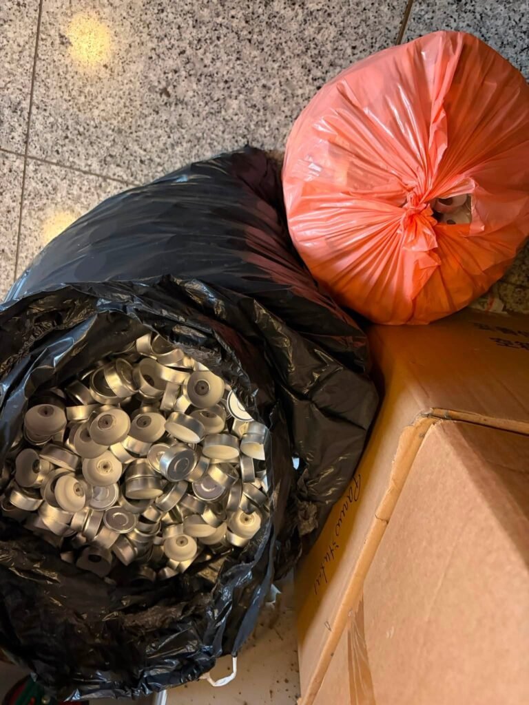 Maišas išdegusių skridininių žvakių indelių ir dar vienas pilnas maišas ant grindų – parafino ir aliuminio žaliava apkasų žvakėms