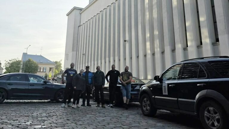 Šeši savanoriai stovi prie automobilių priešais Kauno Kristaus Prisikėlimo baziliką ankstų rugsėjo 8-osios rytą, dešinėje – Kia Sorento su „Visi išvien“ lipduku