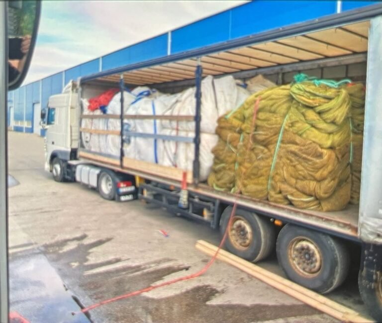Sunkvežimis su atidengtu kėbulu, pakrautas didmaišiais ir žvejybinių tinklų ryšuliais, prie sandėlio