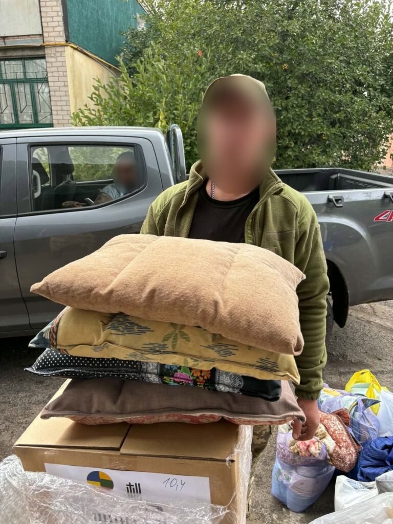 Ukrainos karys laiko šūsnį pagalvių virš dėžių su humanitarine pagalba; veidas uždengtas