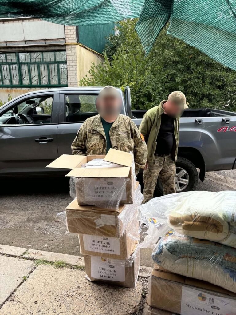 Du Ukrainos kariai prie pikapo su dėžėmis patalynės ligoninei; veidai uždengti