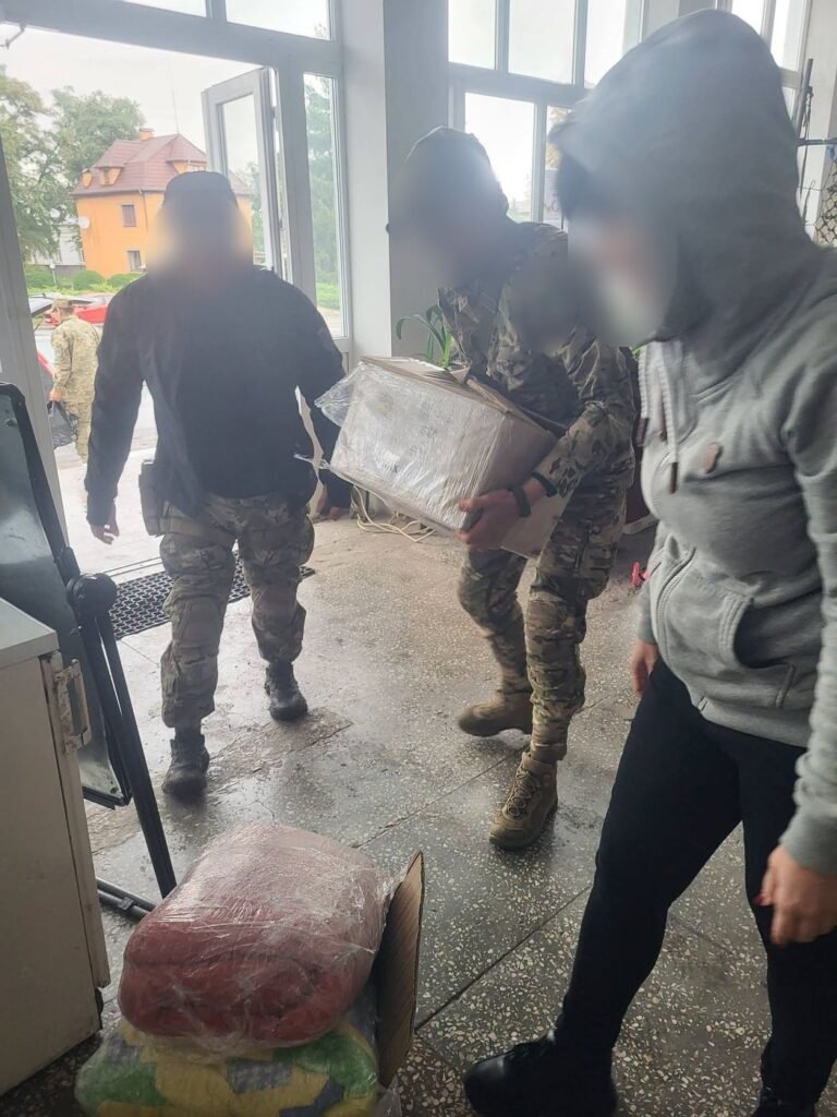 Du Ukrainos kariai ir savanorė neša dėžes ir ryšulius su parama pro stiklines duris; veidai uždengti