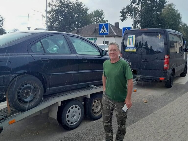 Vairuotojas lietuje stovi prie priekabos su juodu automobiliu, ją tempia fondo mikroautobusas su „Visi išvien“ ženklu