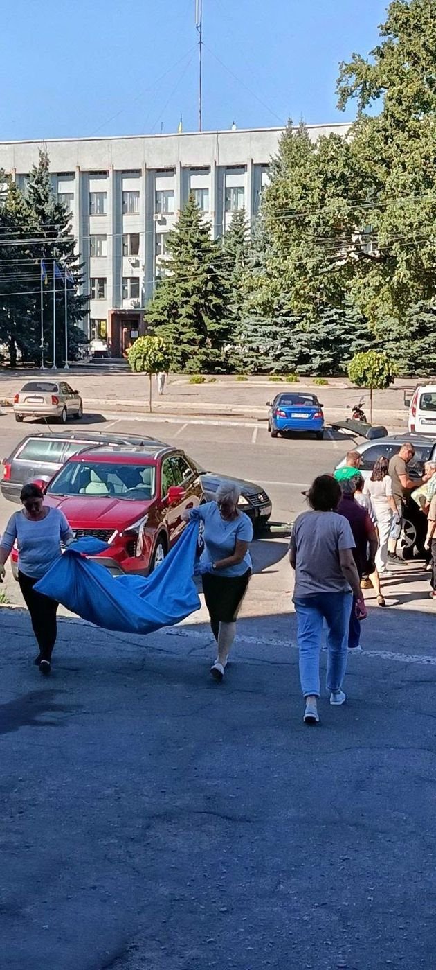 Dvi moterys neša didelį mėlyną ryšulį su parama, už jų – automobiliai Pyriatyno aikštėje