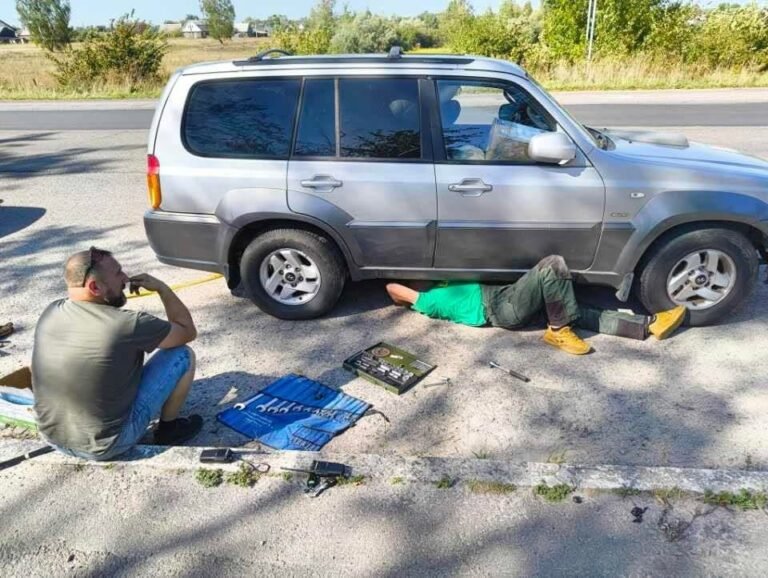 Pakelėje vienas vairuotojas guli po sidabriniu visureigiu ir jį taiso, kitas sėdi šalia prie išdėliotų įrankių