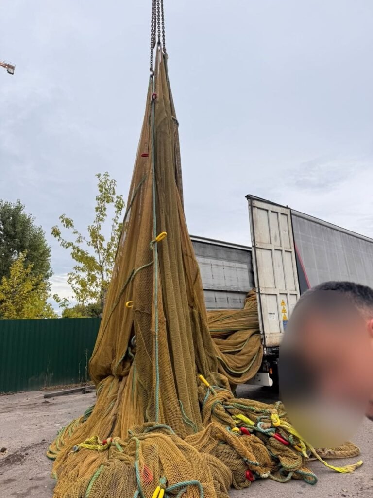 Ant krano kablio pakibęs aukštas gelsvų jūrinių tinklų ryšulys, apačioje – tinklų krūva