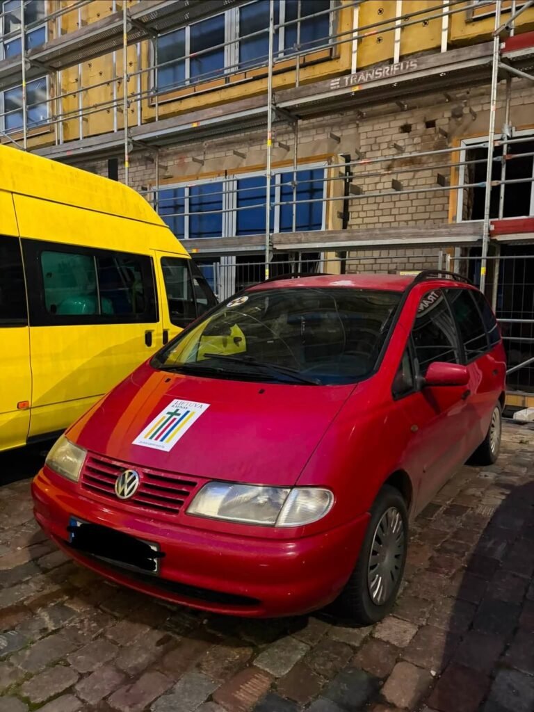 Raudonas VW Sharan su „Visi išvien“ ženklu ant kapoto bazilikos kieme, šalia – geltonas mikroautobusas